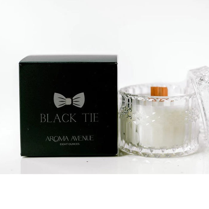 Black box with bow tie logo next to a crystal glass candle jar with a wooden wick