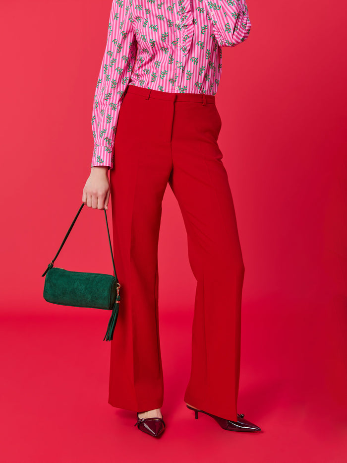 Person wearing a pink striped shirt with green leaf print and red trousers holding a green suede handbag