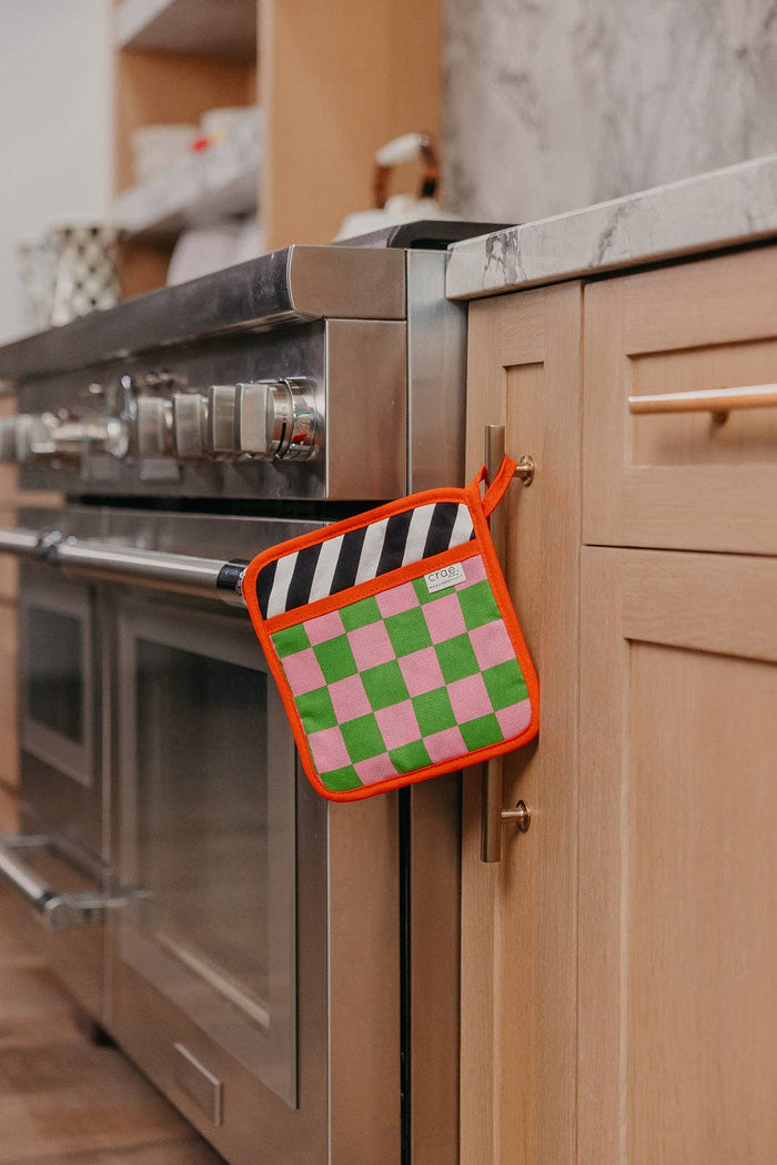 Pink and green checkered pot holder with black and white striped trim hanging on a kitchen cabinet handle