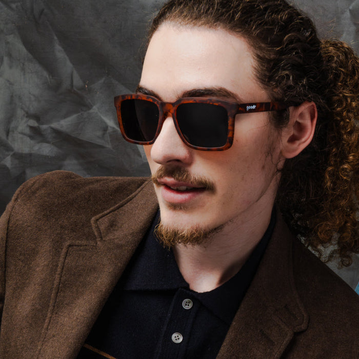 Man in a brown jacket wearing tortoiseshell square sunglasses